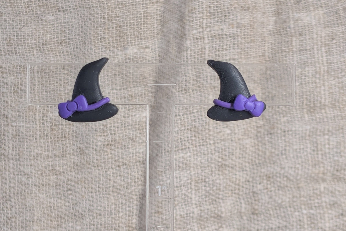 A pair of small witch hat stud earrings in black with purple bows, displayed on a textured beige linen fabric background.