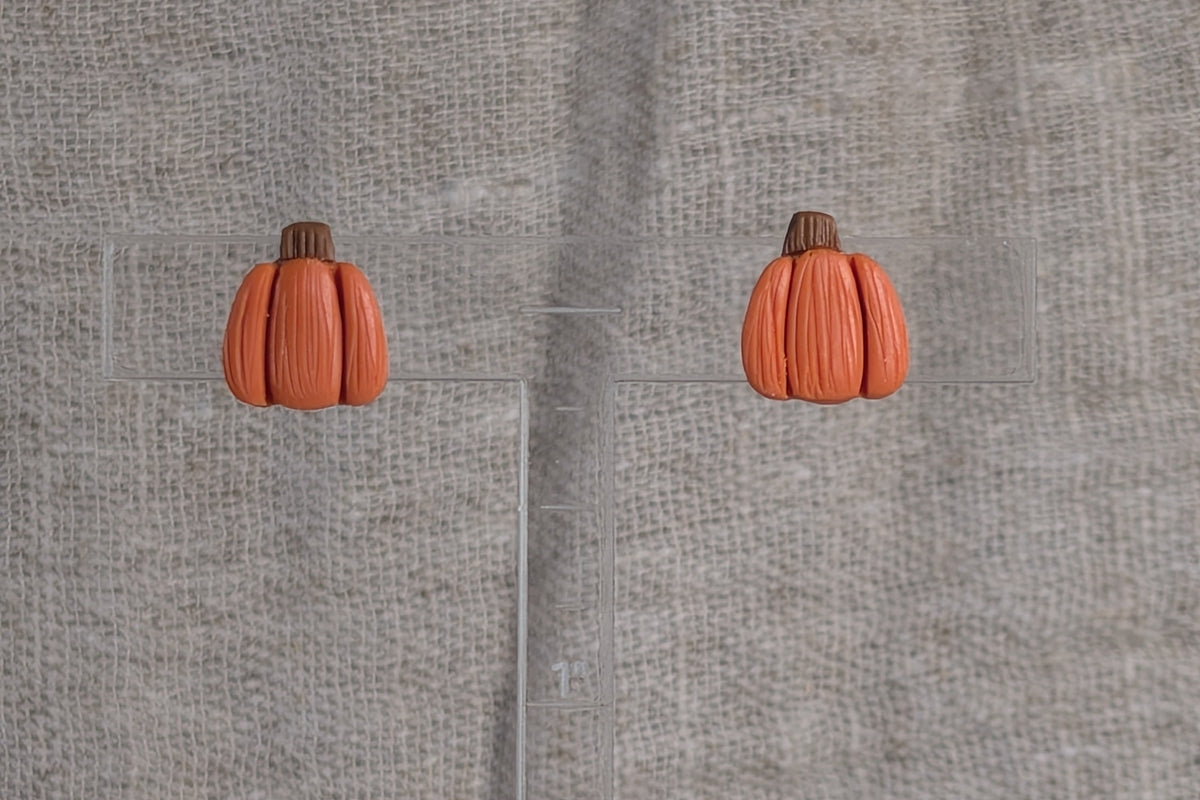 A pair of small orange pumpkin shaped earrings with brown stems and a light texture, displayed on a clear acrylic ruler against a textured beige fabric background.