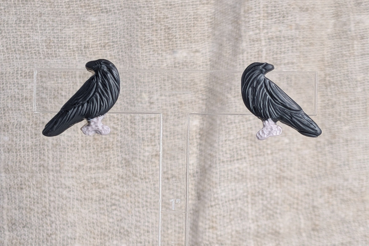 A pair of black raven stud earrings with pale grey feet, displayed on clear acrylic stands against a textured beige linen background.