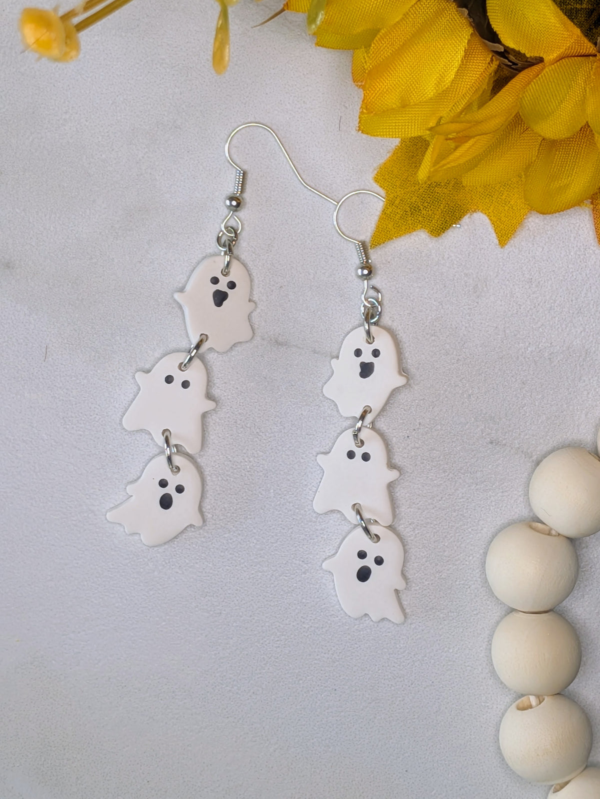 A pair of dangling ghost earrings with white ghost charms and black facial features, hanging from silver hooks on a light background with yellow flowers and white beads as props.