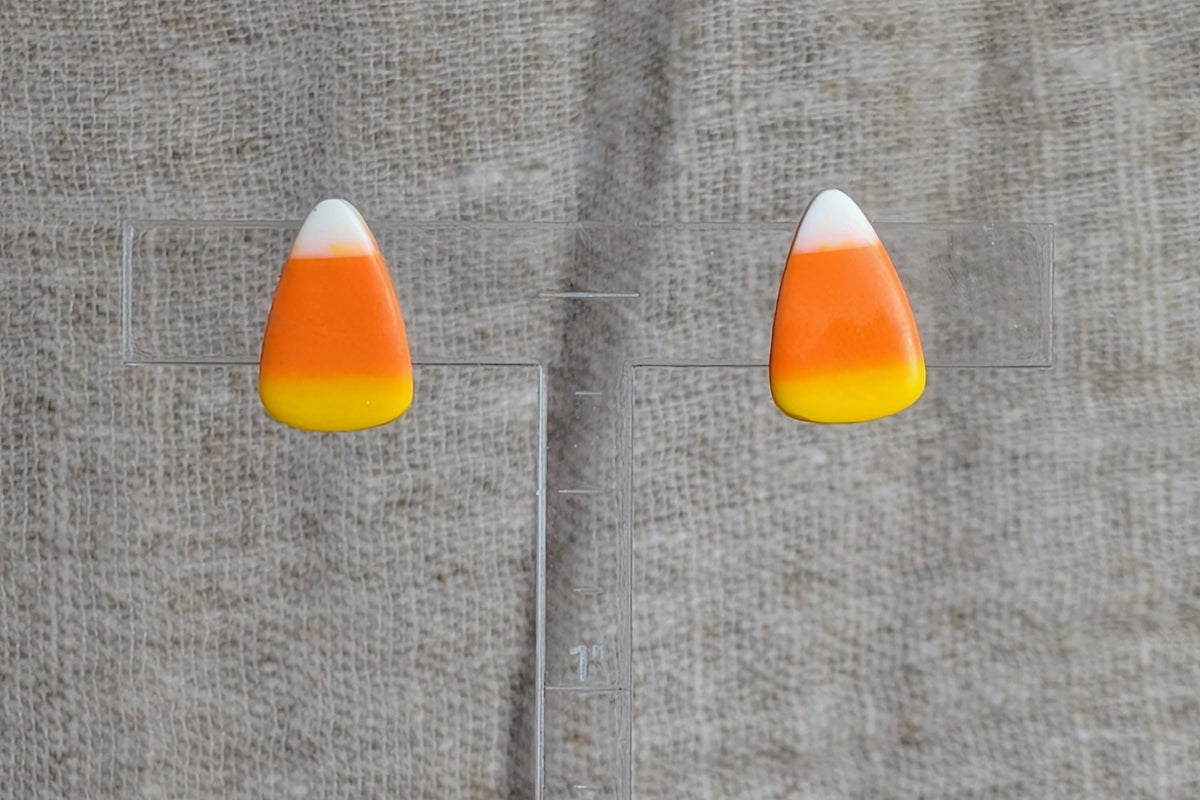 A pair of candy corn stud earrings with white, orange, and yellow sections, displayed on a clear acrylic block against a textured beige linen background.