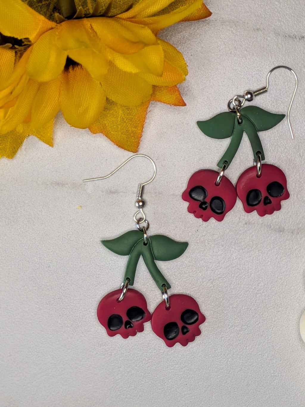 Cherry blossom skull drop earrings with green leaves and red cherry charms featuring black skull details, displayed on a light textured surface with a yellow flower prop.