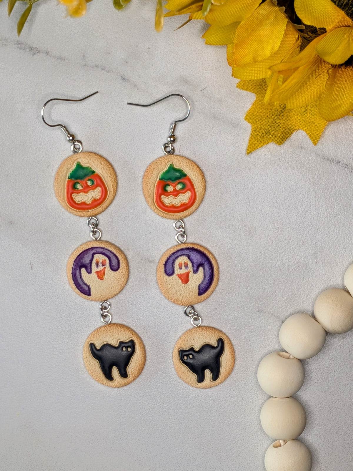 A pair of dangle earrings with silver hooks and circular cookie charms featuring Halloween designs: a jack-o'-lantern on top, a purple ghost in the middle, and a black cat on the bottom, displayed on a light marble surface with yellow flowers and white beads as props.