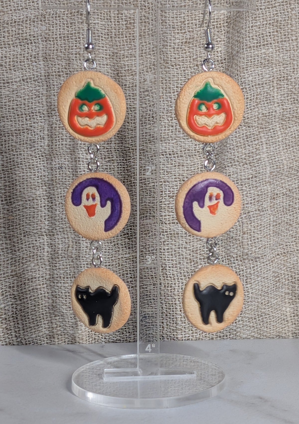 A pair of handmade dangle earrings displayed on a clear acrylic stand against a textured beige fabric backdrop. Each earring features three circular cookie charms decorated with Halloween illustrations: a green and orange pumpkin, a purple ghost, and a black cat.