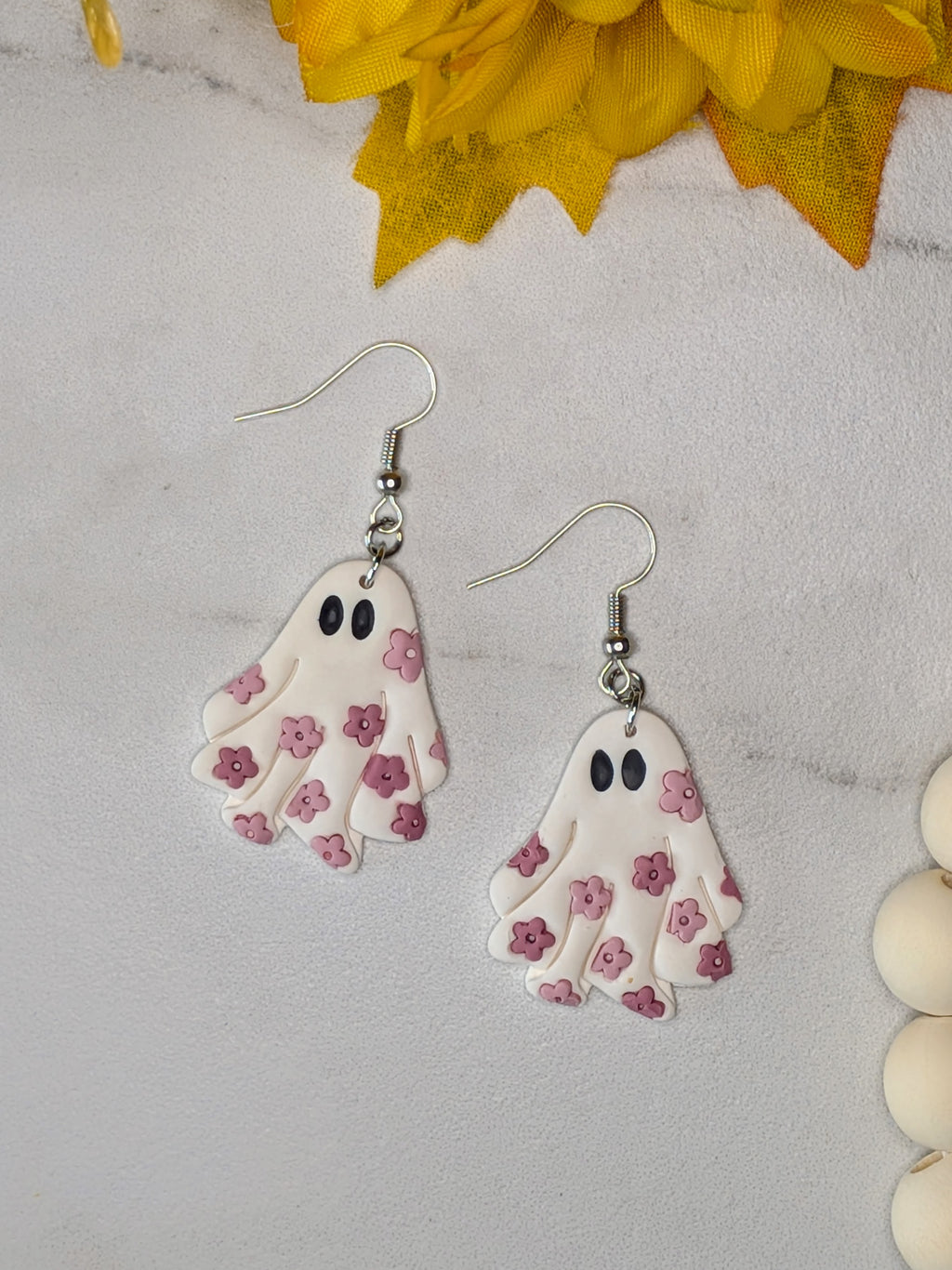 A pair of dangling ghost-shaped earrings with pink flower patterns and black eyes, hanging from silver fishhook wires on a light gray surface with yellow decorative leaves and white beads nearby.