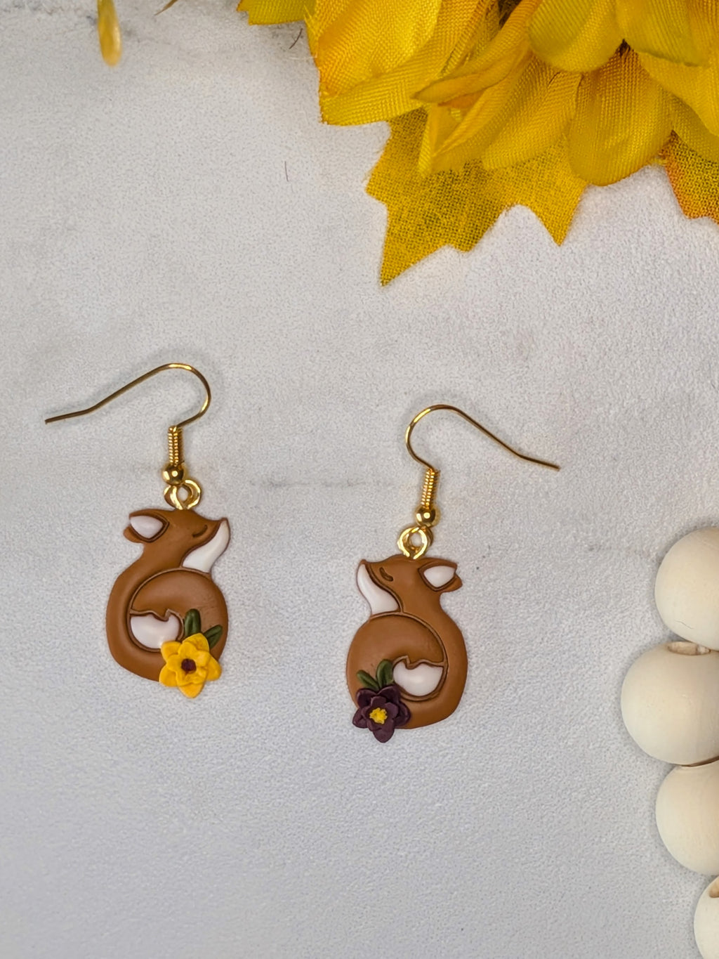 A pair of dangling earrings with terracotta colored earrings showing curled foxes with white accents and small yellow and purple flowers near the bottoms, displayed on a light textured surface with yellow leaves and white beads as props.