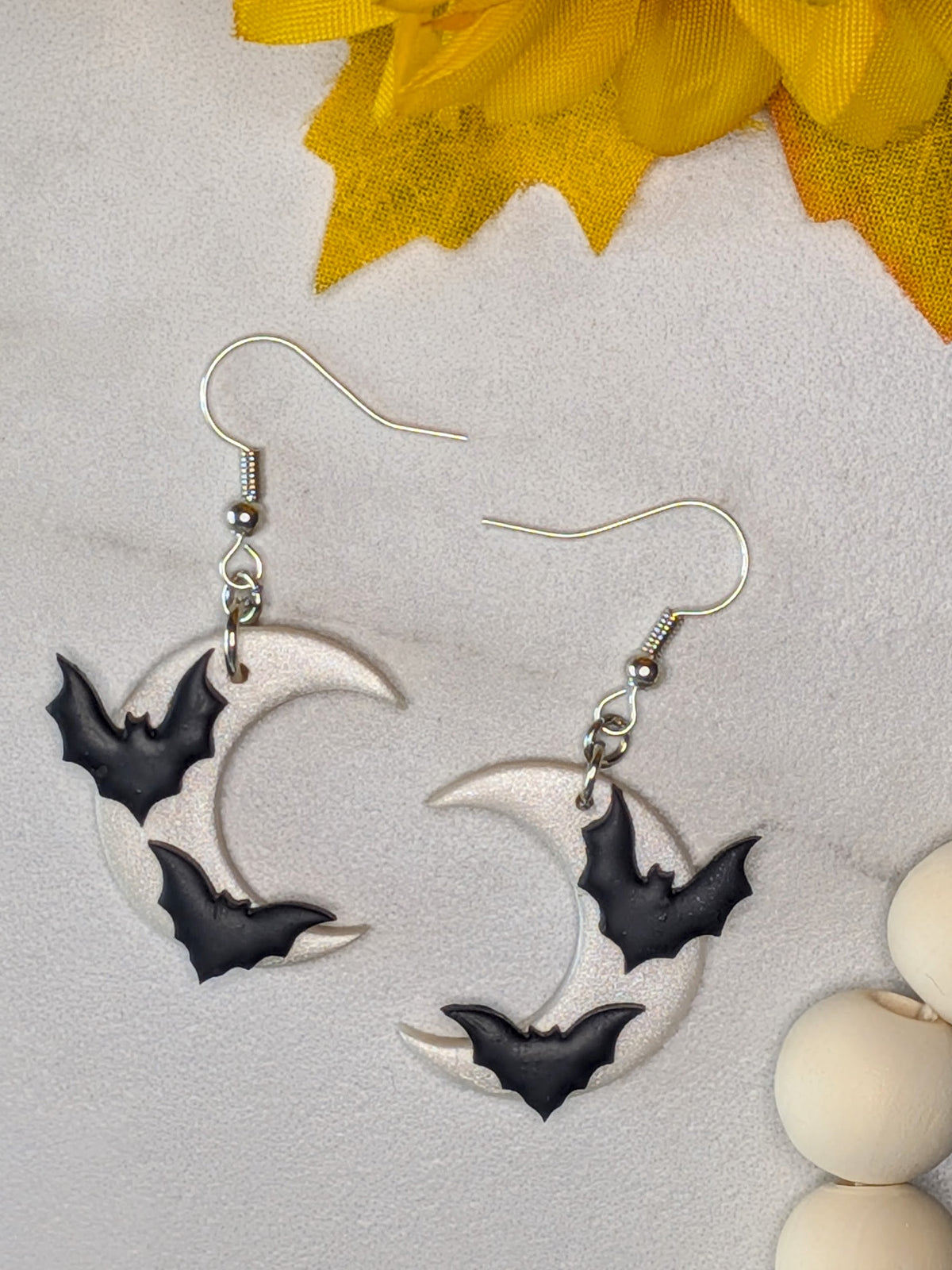 A pair of white-silver fishhook earrings with crescent moon and bat charms, displayed on a light textured surface with yellow and white decorative props.