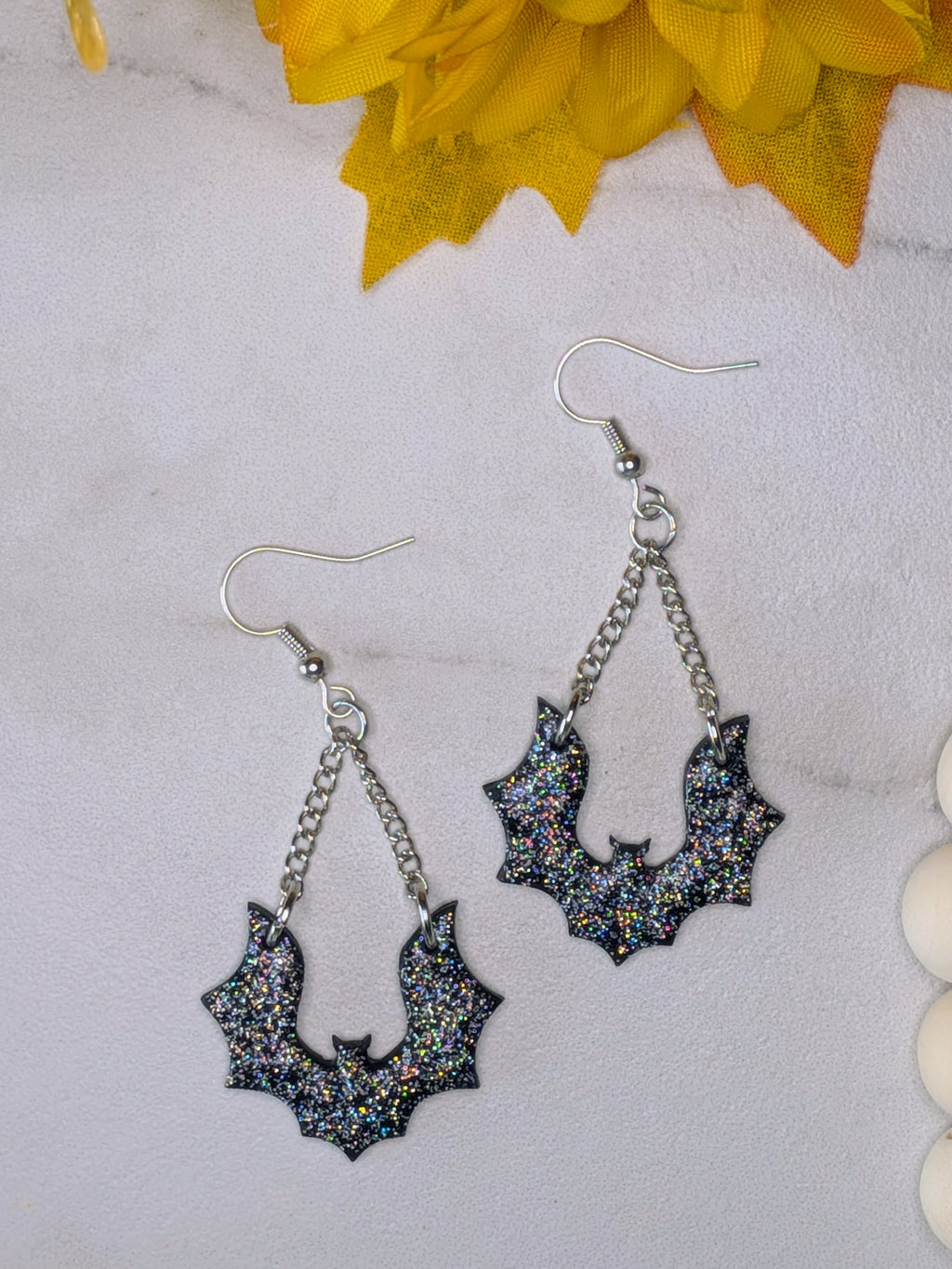 A pair of dangling bat-shaped earrings with silver-tone hooks and chains, covered in black glitter with multicolor iridescent flecks, displayed on a light marble-like surface with yellow flowers in the corner.