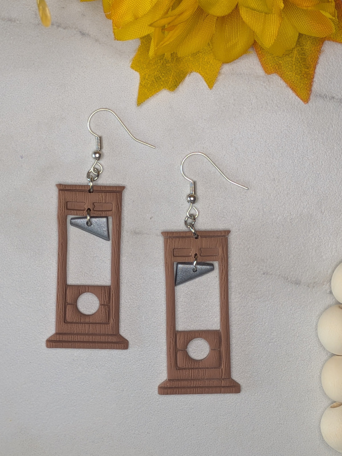 A pair of dangling earrings shaped like wooden guillotine blocks, with silver fishhook wires, displayed on a light marble-like surface with yellow flowers and white beads as props.