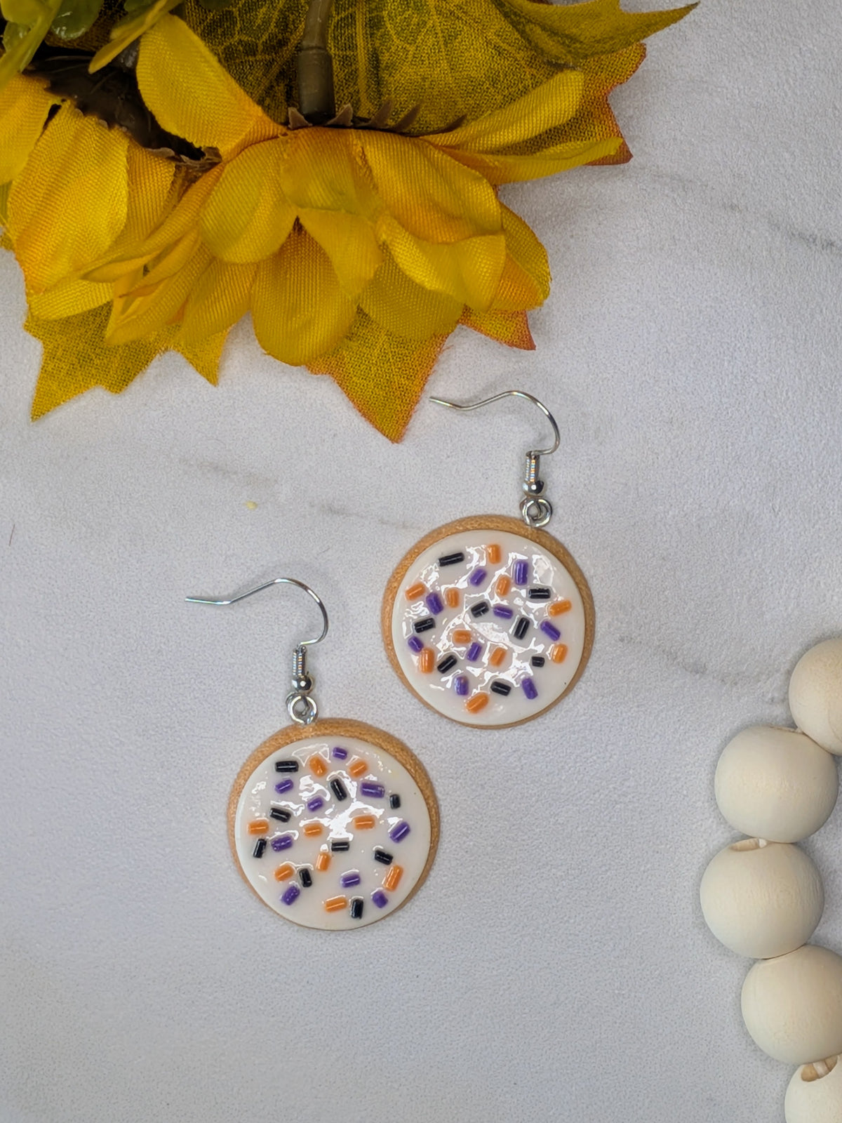 A pair of sugar cookie shaped dangle earrings with silver fishhook wires, decorated with white frosting and purple, black, and orange sprinkles, displayed on a light marble surface with yellow autumn leaves and white beads as props.