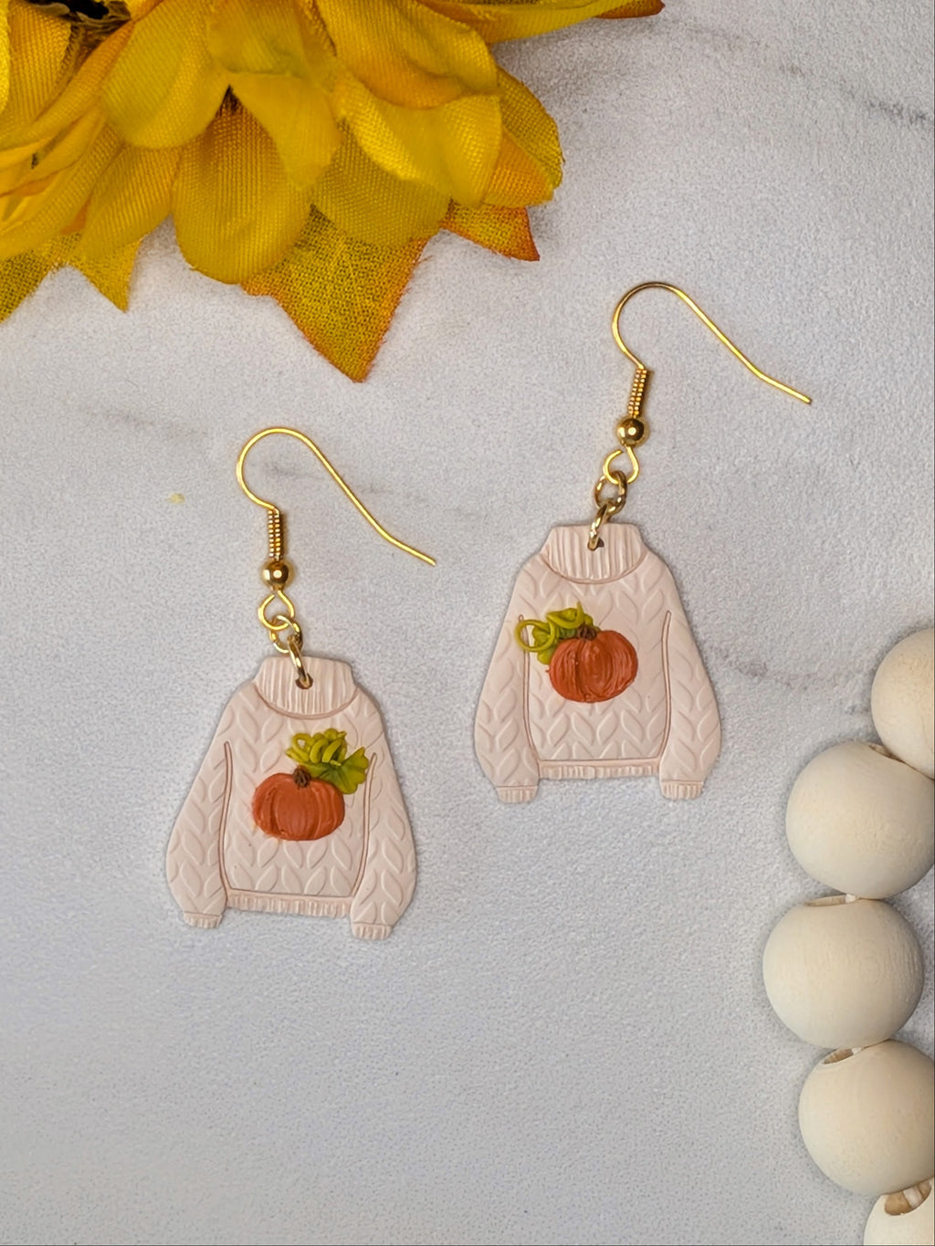 A pair of dangling earrings shaped like cream-colored cable-knit sweaters with orange pumpkin patches backed with green leaves and curly vines on the front, hanging from gold fishhook wires. The earrings are displayed on a light gray marble surface next to yellow autumn leaves and white wooden beads.